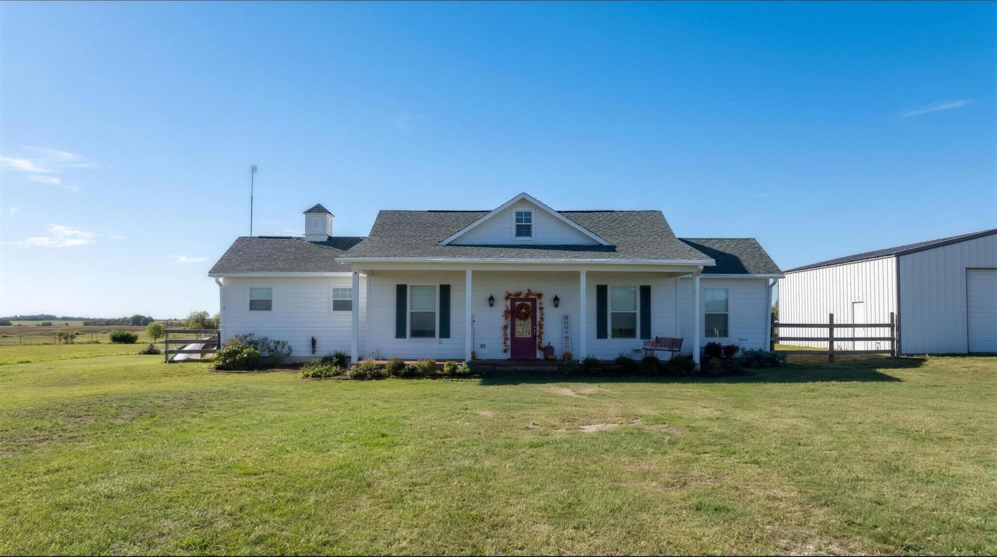 The white farmhouse with green shutters, a red front door, and a rooftop cupola, its metal barn and open pasture beside it under a clear blue sky