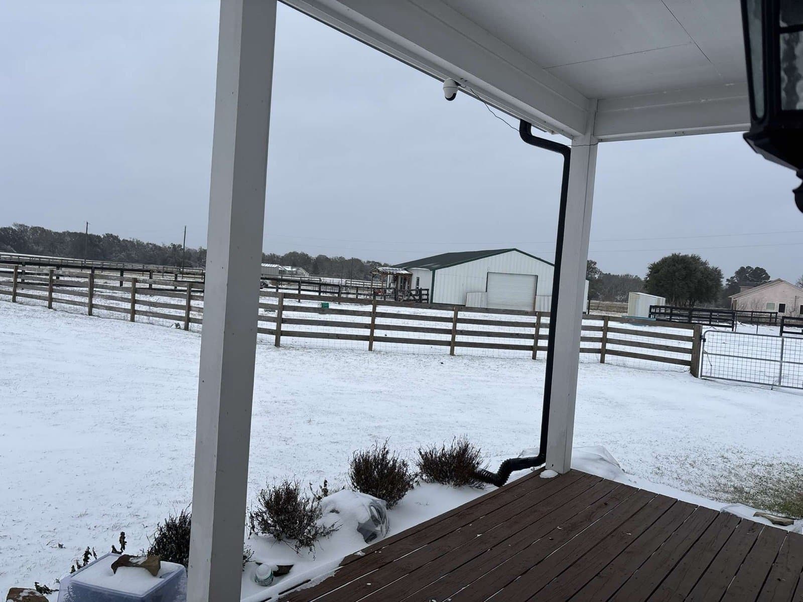 Snow blanketing the pasture, fence line, and metal barn, seen from the covered porch