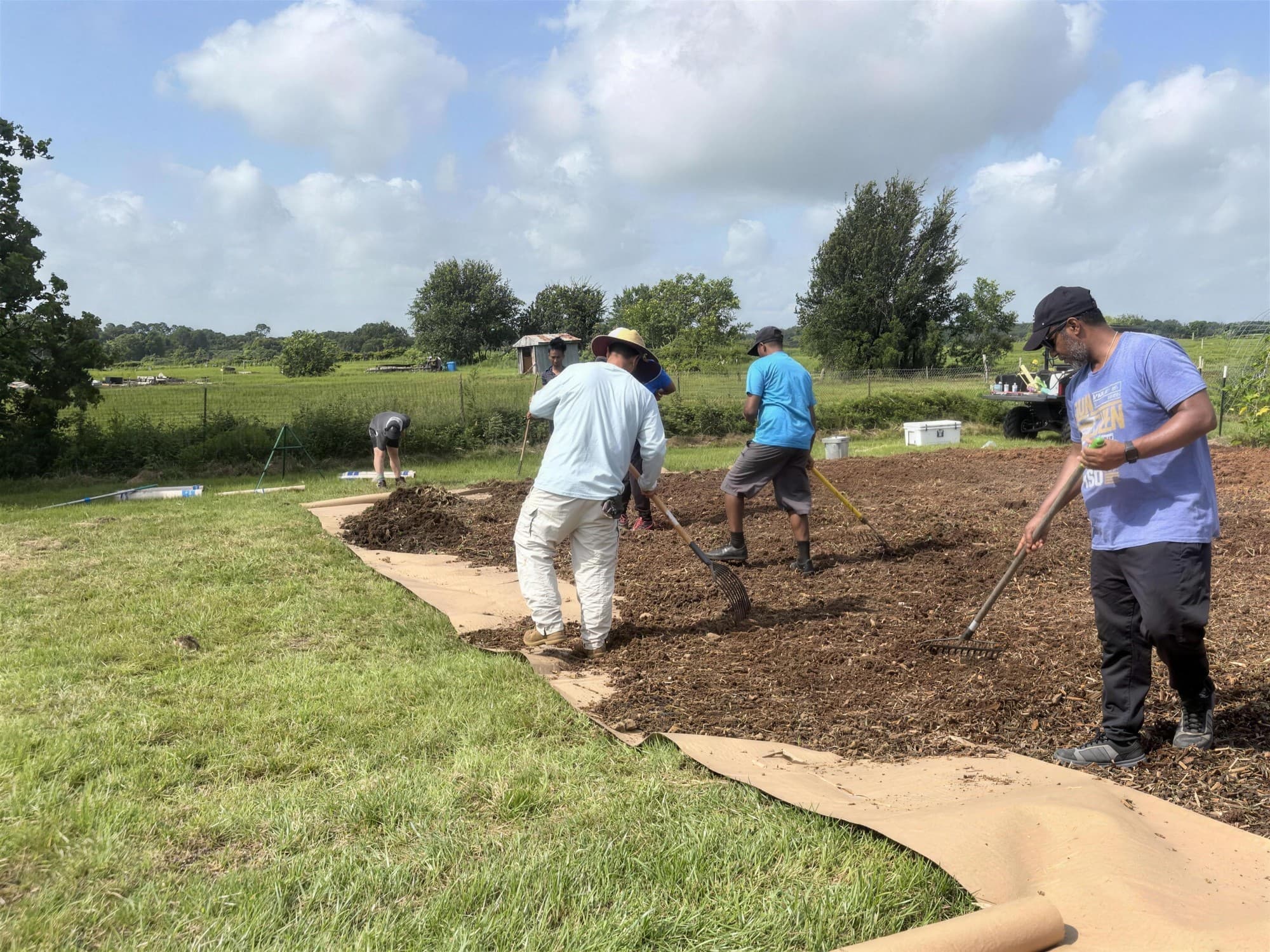 A small group works together turning and raking soil in a garden bed on the farm, with open pasture behind them.