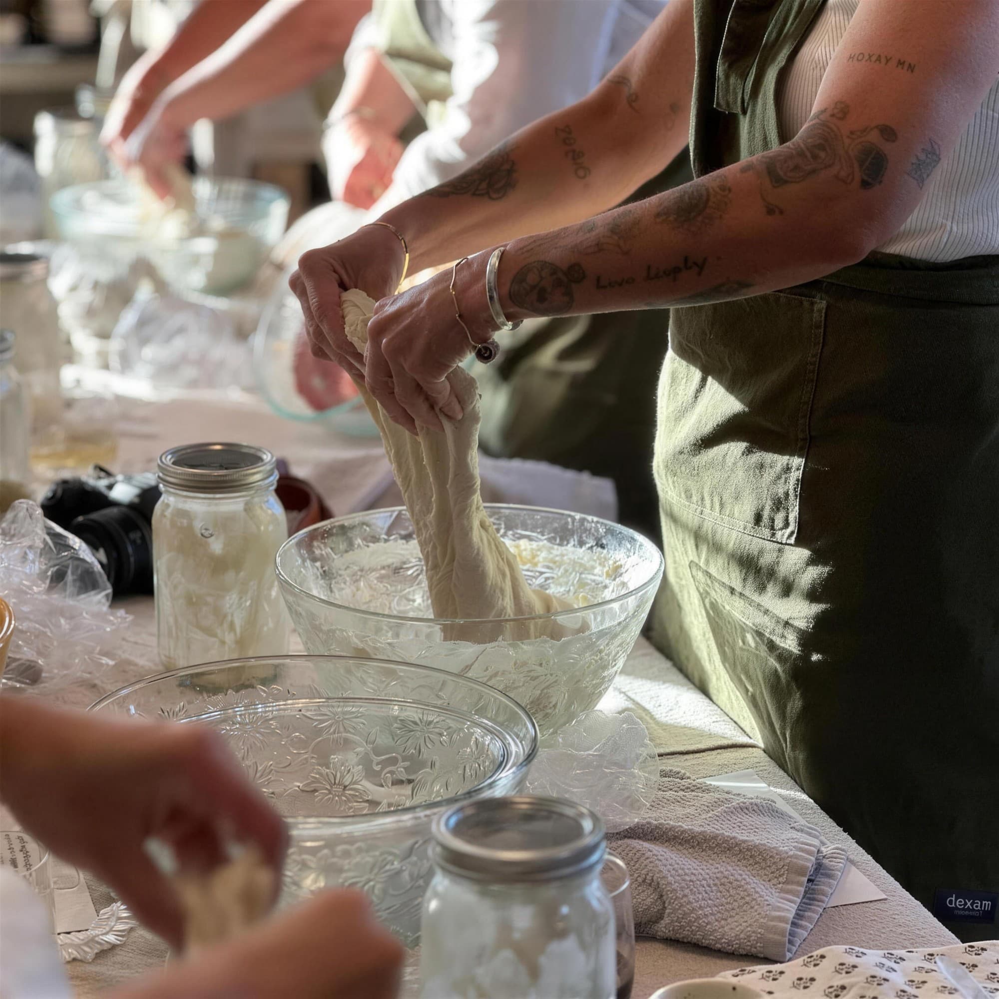 A person in an apron stretching a mass of sourdough dough over a glass bowl, with jars of starter on the table.