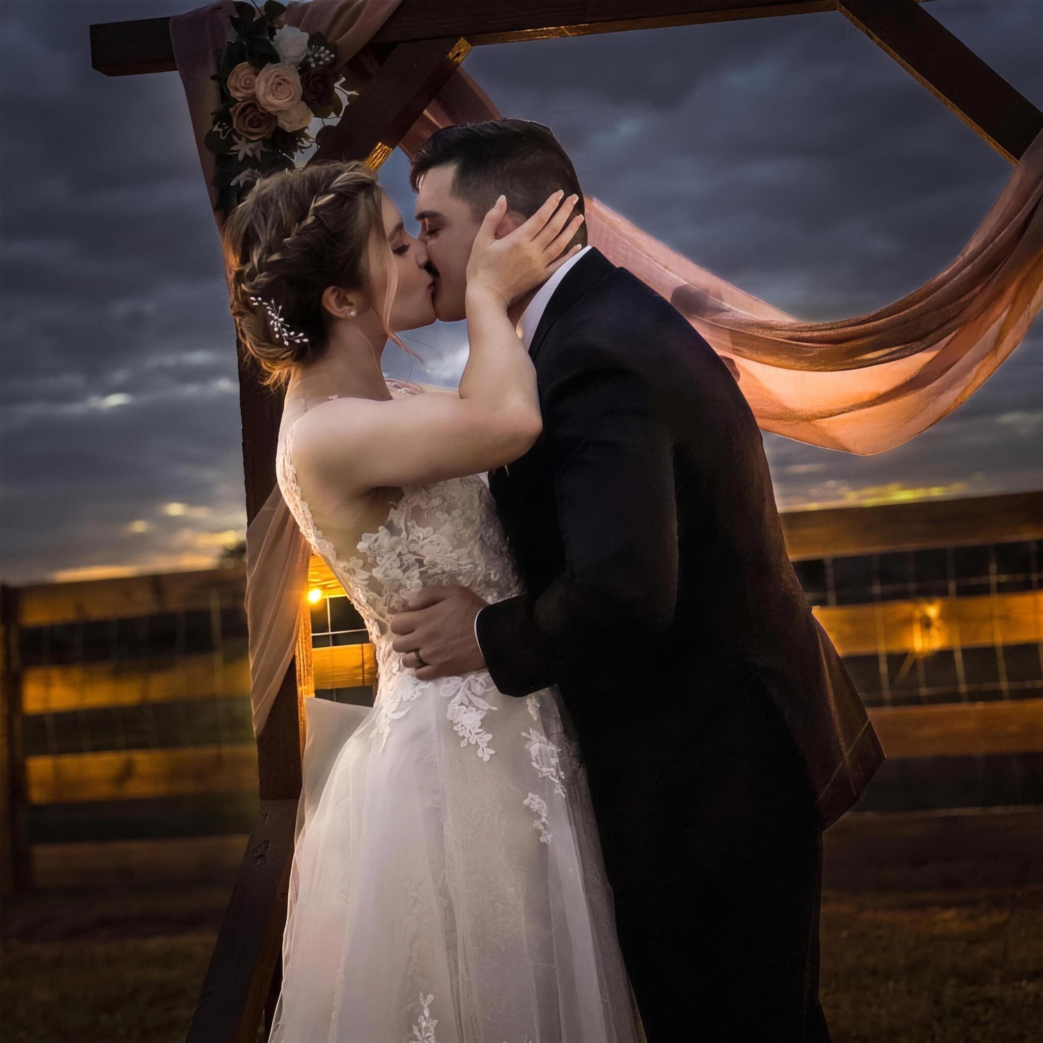 A bride and groom kissing under a wooden wedding arch draped with fabric and flowers as the sun sets behind them