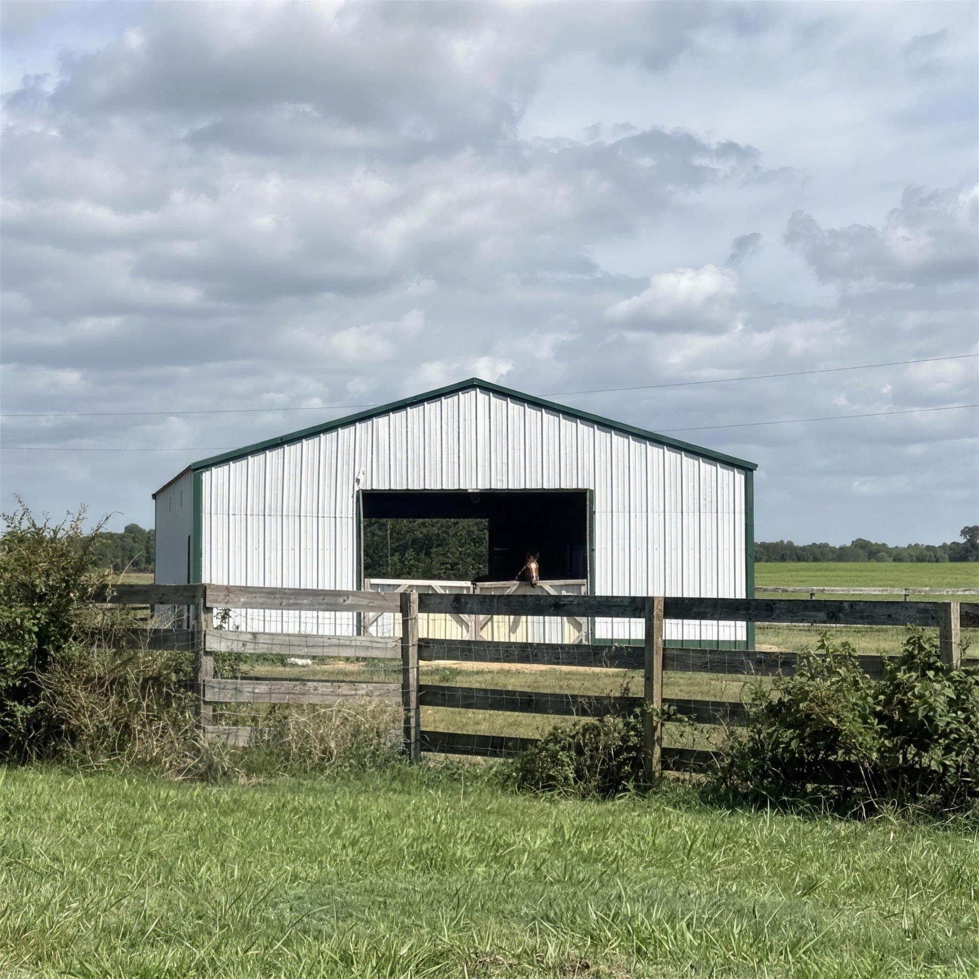 The big white horse barn with green trim under a wide cloudy sky, a horse looking out from the open bay behind the wooden paddock fence