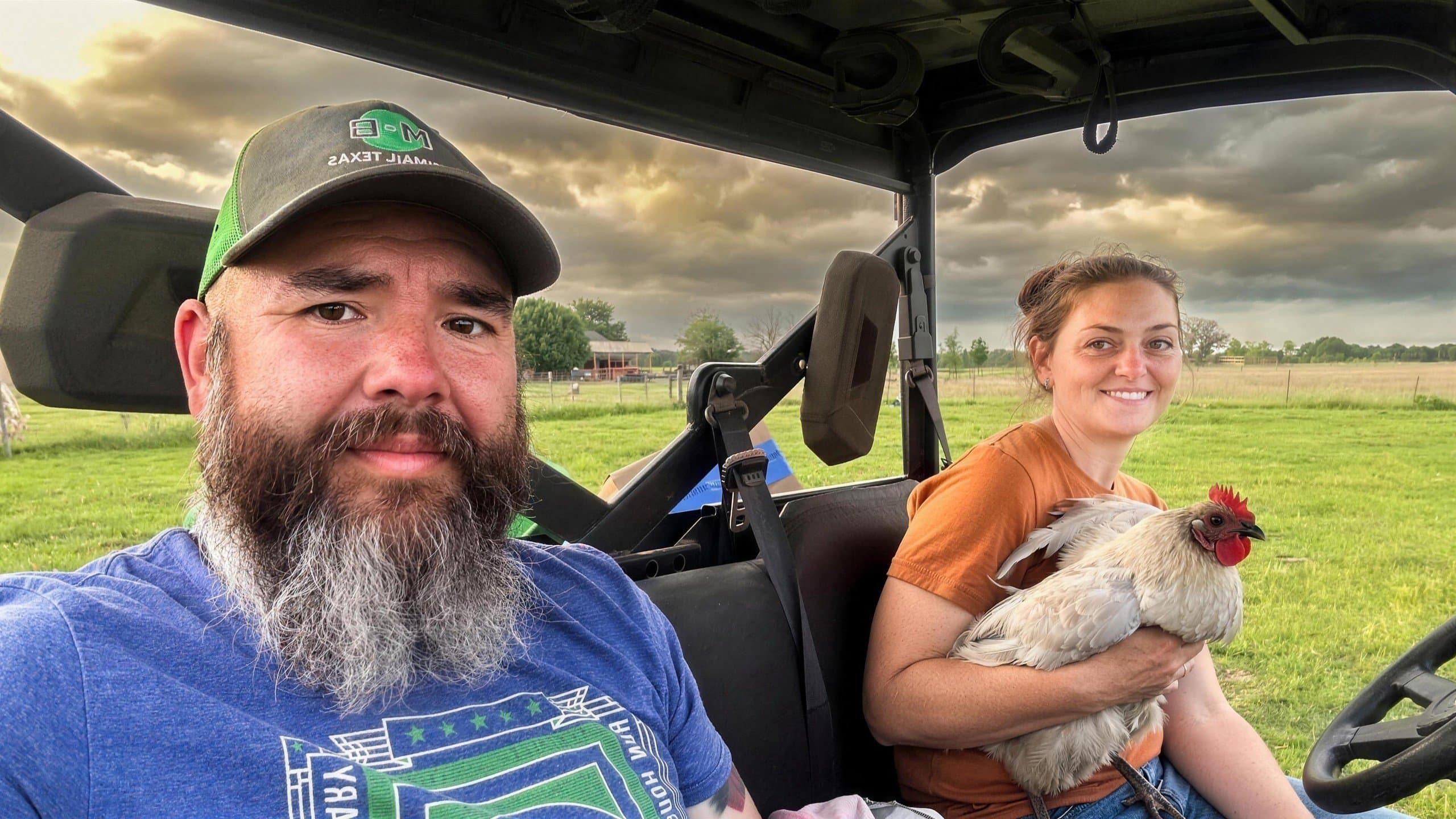 Joe and Tori Bell riding in their farm UTV, Tori holding a speckled cream hen, green pasture and storm clouds behind them
