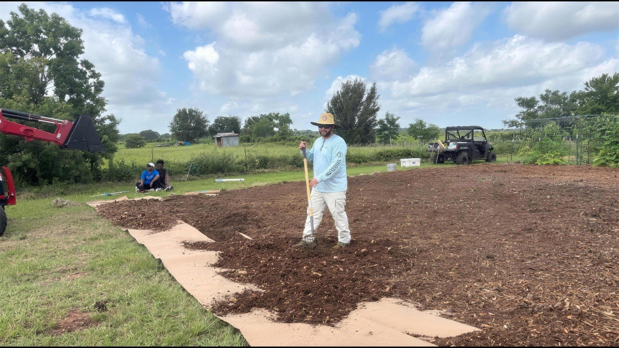A volunteer in a straw hat raking soil in a freshly prepared garden bed.