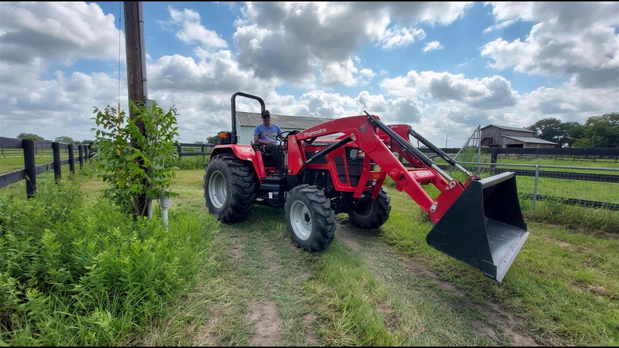 A man on a red tractor with a front loader, parked by the pasture fence with a barn behind.