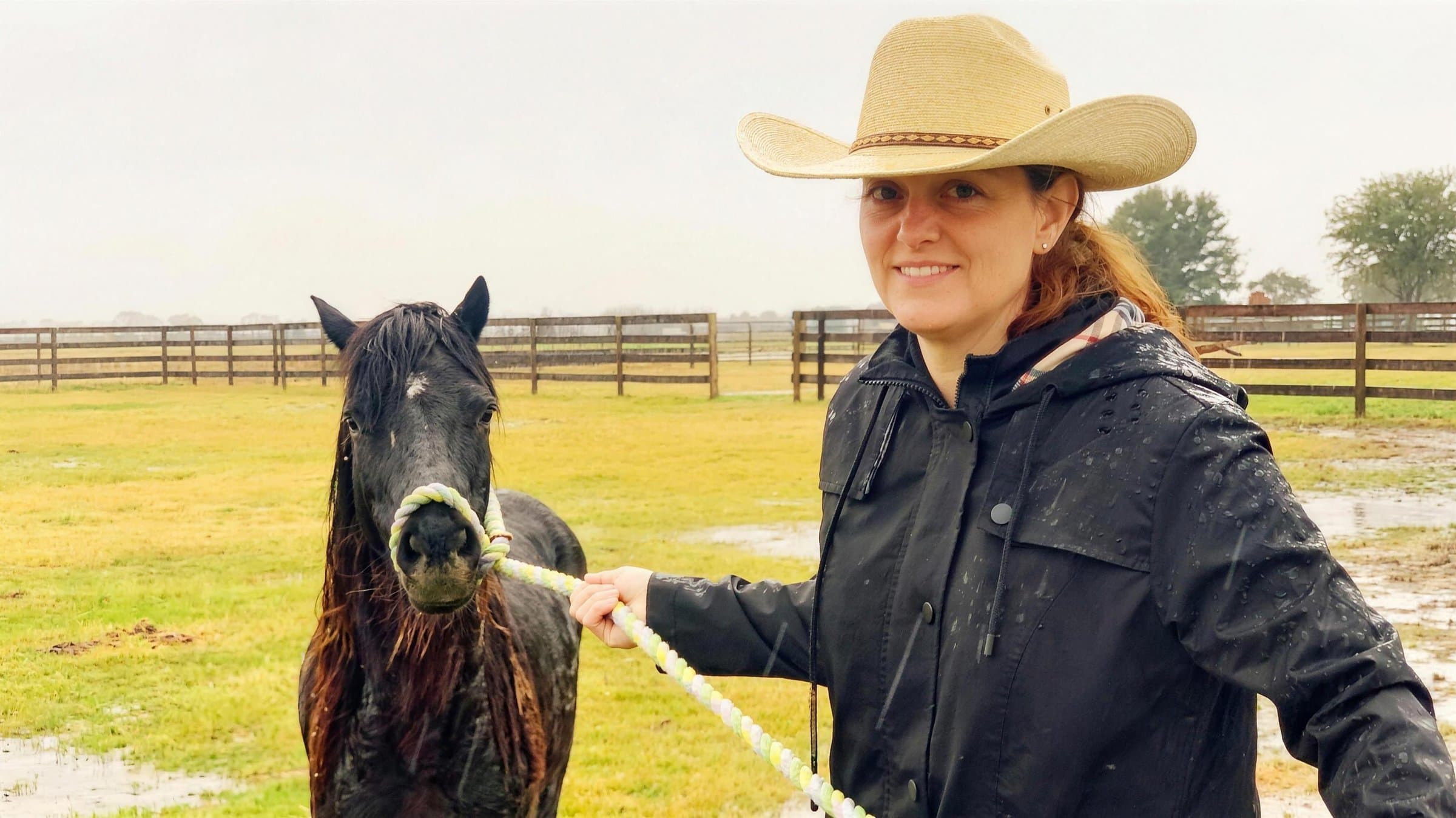 A woman in a straw cowboy hat and black rain jacket leading a wet black horse by a rope halter across a green pasture on a rainy day